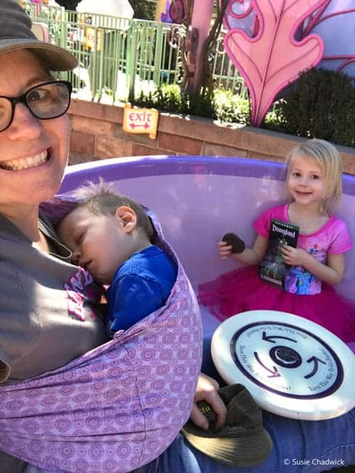 Mom, little girl and baby on a Disney ride. The baby is asleep in a sling the mom is wearing.